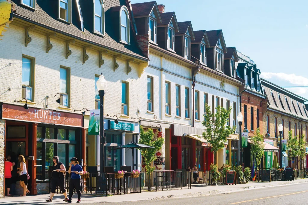 A vibrant street with historic low-rise buildings, colorful storefronts, and people walking along the sidewalk on a sunny day