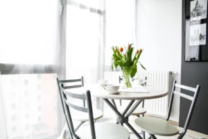 A small table with chairs positioned in front of a window, creating a cozy and inviting atmosphere. On the table, there is a vase with flowers. The entire picture is in black and white, except the flowers, which are green