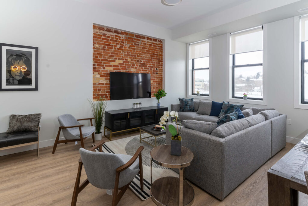 A modern, open-concept living room in a Y-Lofts apartment. It features a large window, a sofa with pillows, and contemporary decor, suggesting a comfortable and stylish urban living space